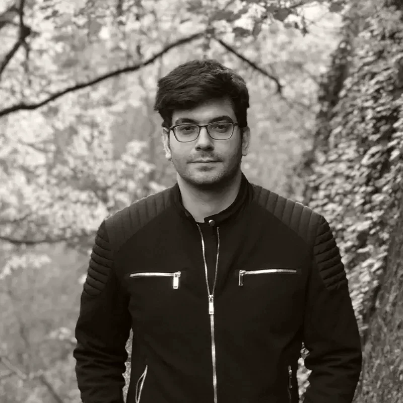 Saleem Najjar, in a dark jacket and glasses, standing outdoors against autumn woodland