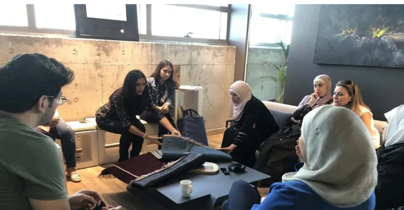 A small group seated around a low table while a mentor holds up a woven textile for the maker sitting opposite her