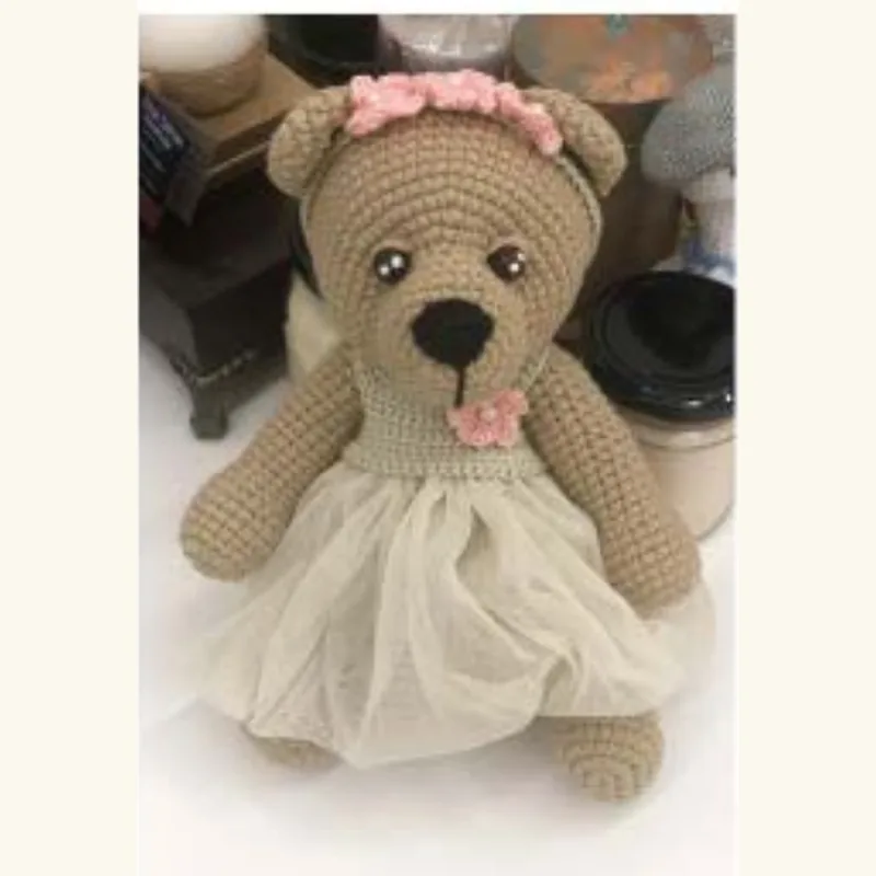 A crocheted bear in a lace dress and dust-coloured yarn, photographed on a shelf